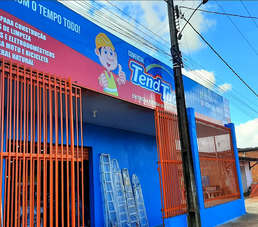 Fachada azul da Comercial Tend Tudo com grades laranja, letreiro com o mascote e escadas expostas na entrada, no Povoado Vila Aparecida em Arapiraca